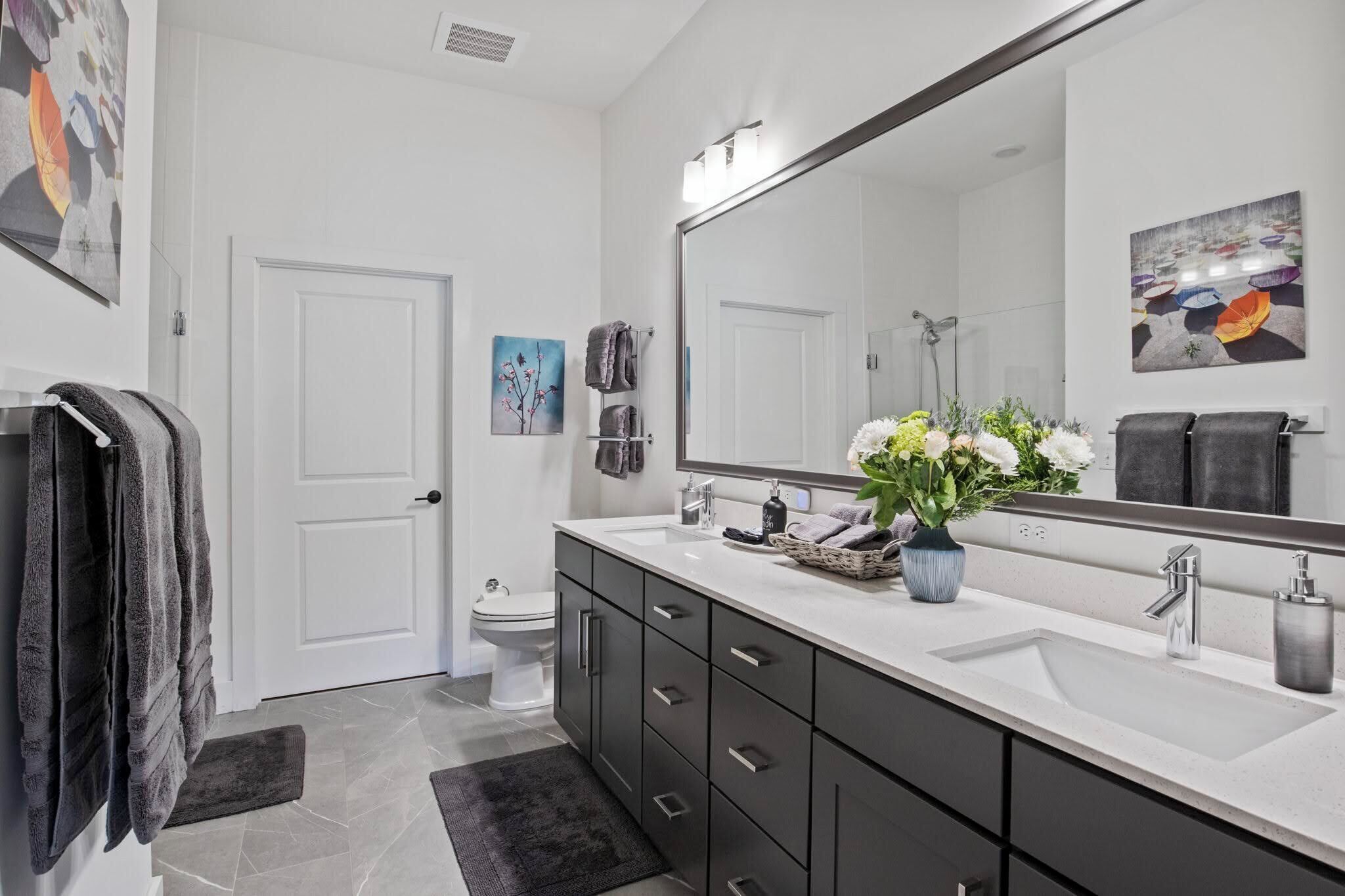 Une salle de bain moderne avec un grand miroir, deux lavabos, des toilettes et une douche. On y trouve des serviettes sur un porte-serviettes, un vase avec des fleurs et des œuvres d’art accrochées aux murs.