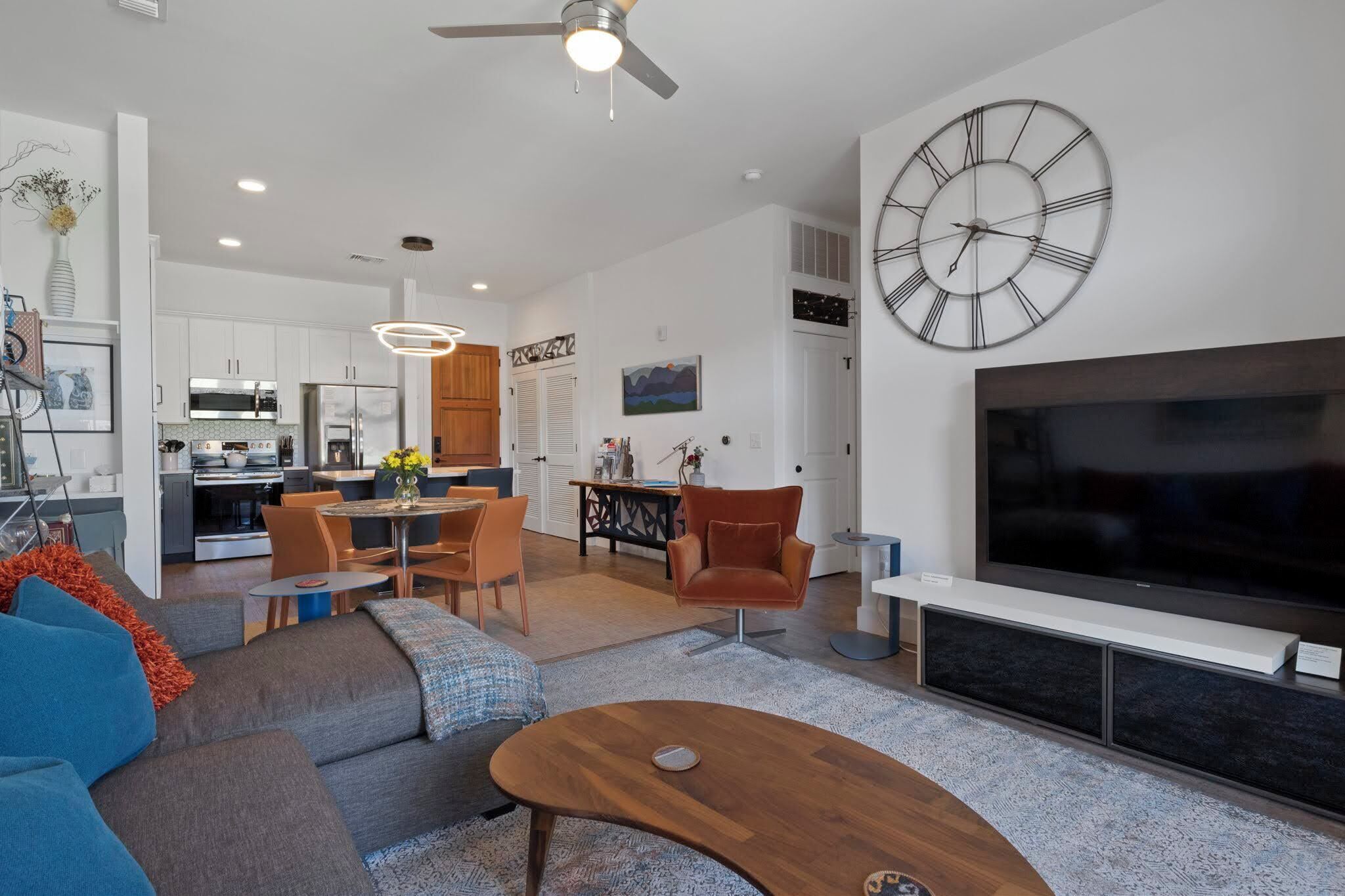 A modern living room with a large flat-screen TV, a dining area with a wooden table and chairs, and a kitchen in the background.
