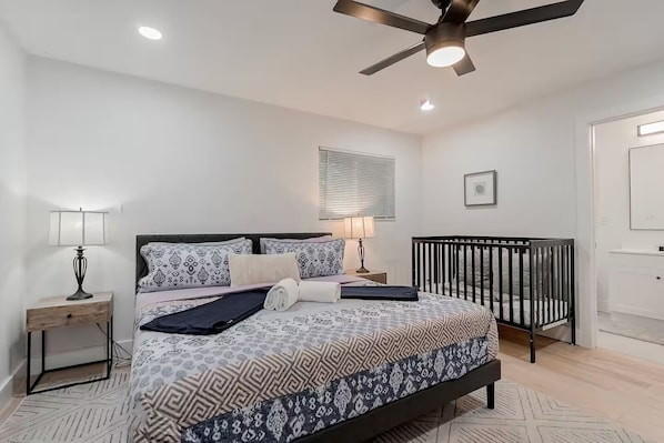 A bedroom with a bed, bedside tables, a crib, a ceiling fan, and a window with blinds.