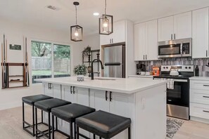 Cocina moderna con armarios blancos, una isla central con taburetes de bar, electrodomésticos de acero inoxidable e iluminación colgante.