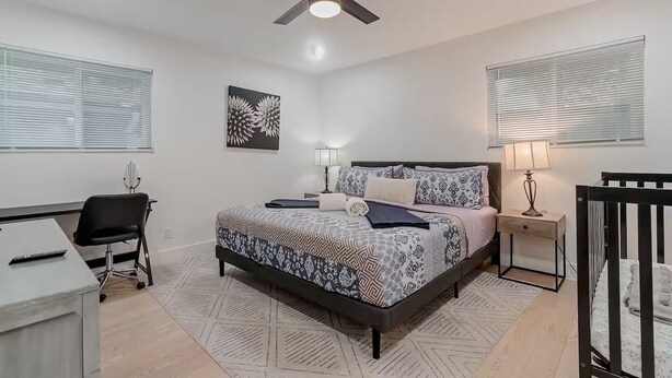 A bedroom with a bed, bedside tables, a desk, a chair, a ceiling fan, and a window with blinds.
