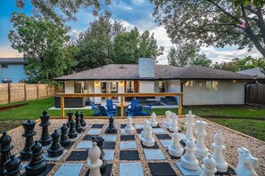 A residential property with a large outdoor chess set, a covered patio area, and a house with a visible chimney. - Spacious 4BR at Austin Hideaway, Firepit, Giant Chess, Hot Tub, Games, Sleeps 12 (Austin)