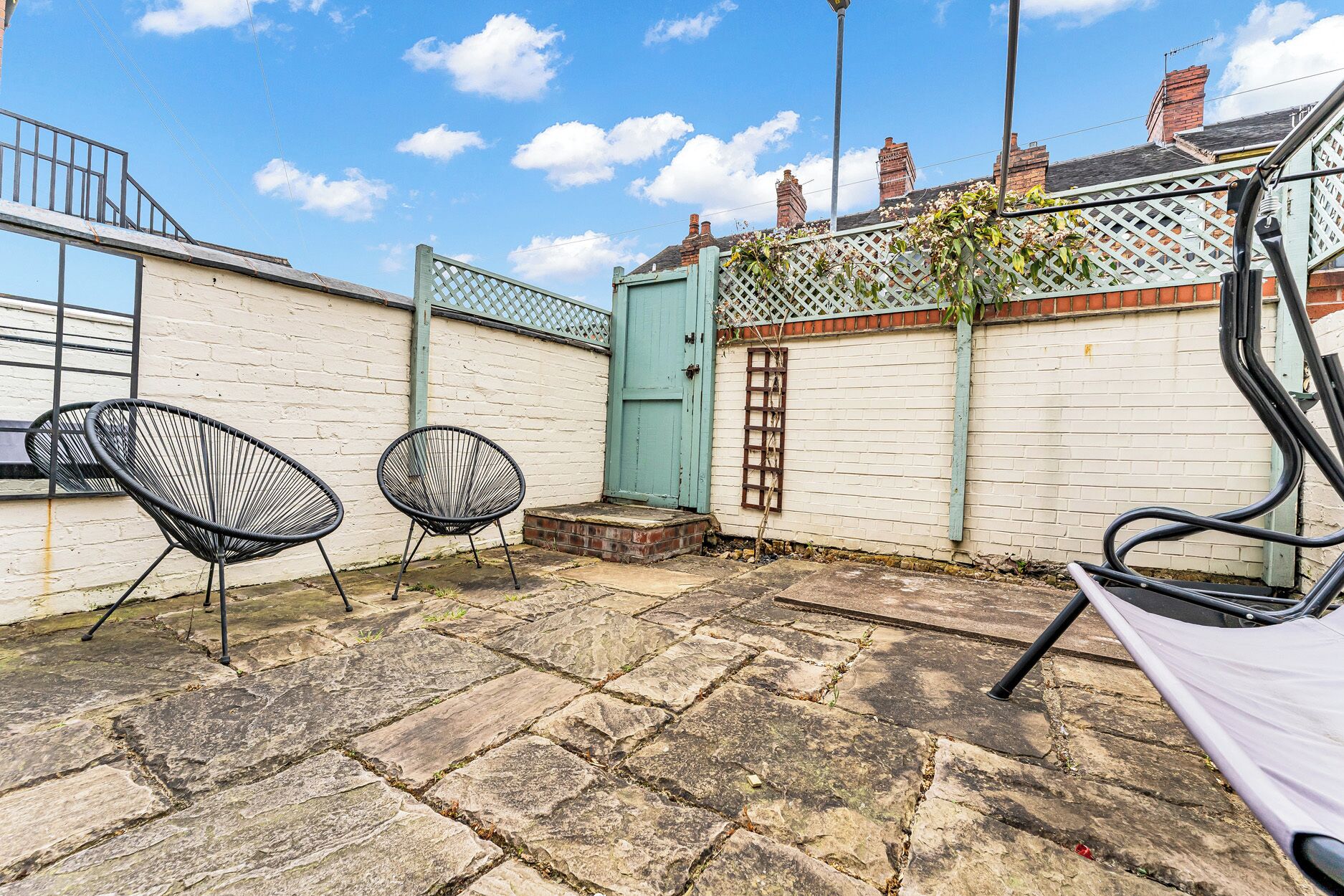 A courtyard with stone paving, two black wireframe chairs, a blue door, and a white fence with climbing plants.