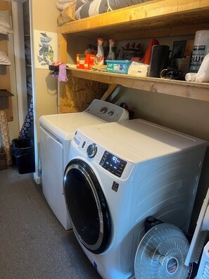 A compact laundry area with a front-loading washer and dryer, a small shelf with cleaning supplies, and a fan. - Beautifully renovated 2-bedroom/1 bath apt. Parking and Laundry on site. (Burlington)