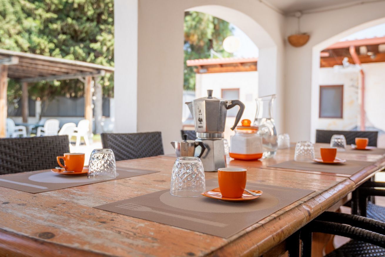 Un tavolo di legno con tazze arancioni e una caffettiera, in una zona pranzo all'aperto con sedie e una pergola.