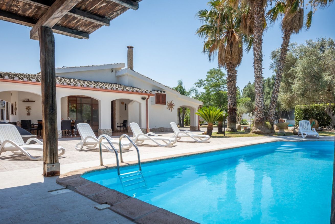 Un área de piscina con sillones de descanso, una pérgola y palmeras.