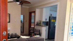 A living room with a grey sofa, a dining area with a table and chairs, a kitchen with cabinets and appliances, and a ceiling fan. - Deluxe Unique 2BR/2BA Apartment – Stylish, Modern at San Cristobal City, DR. (San Cristóbal)