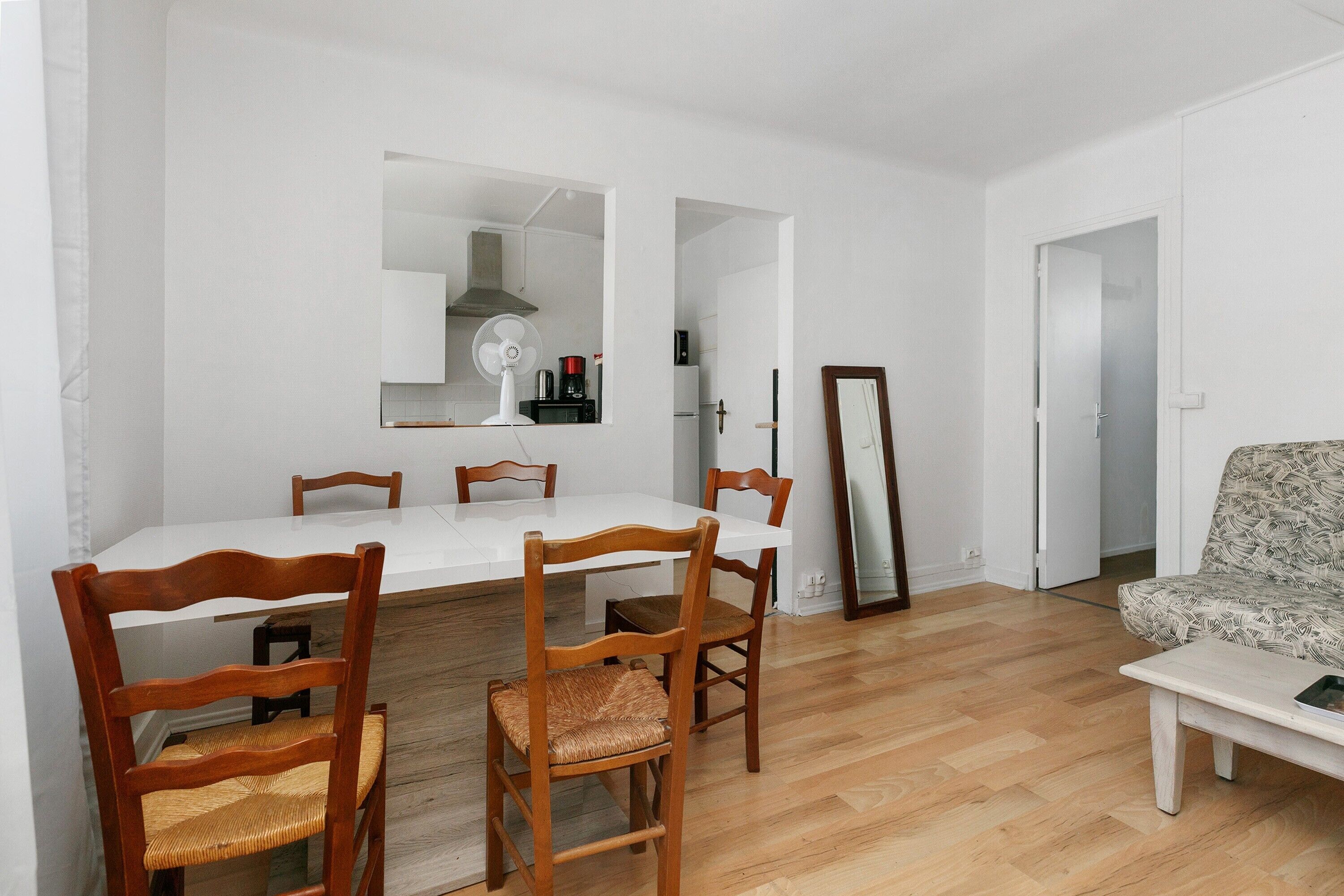 A dining area with a white table and wooden chairs, a kitchen visible through an open doorway, and a sofa in the adjacent living room.