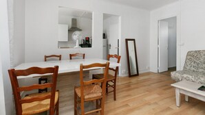 Un'area pranzo con tavolo bianco e sedie di legno, una cucina visibile attraverso un'apertura, e un divano nella sala da pranzo adiacente.