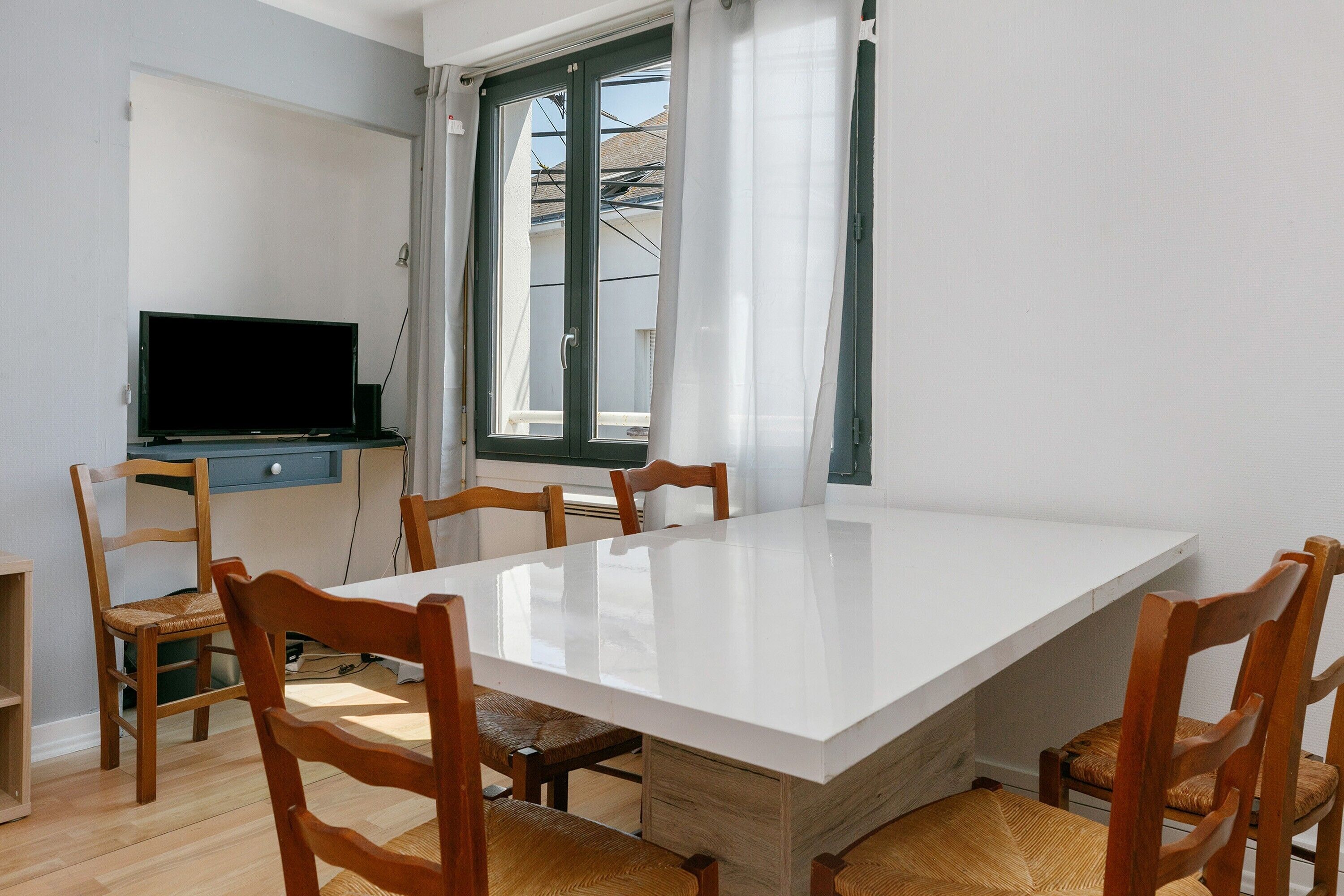 Un comedor con una mesa blanca, sillas de madera, un televisor sobre un mueble y una ventana con cortinas.