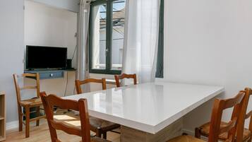 Un comedor con una mesa blanca, sillas de madera, un televisor sobre un mueble y una ventana con cortinas.
