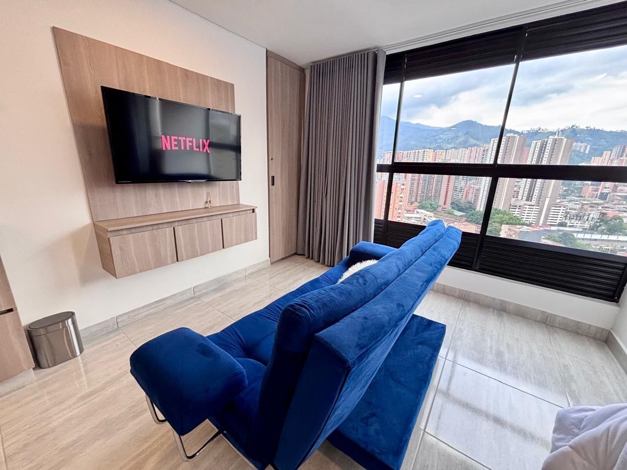A modern living room with a long blue sofa, a mounted TV, and a large window with a city view.
