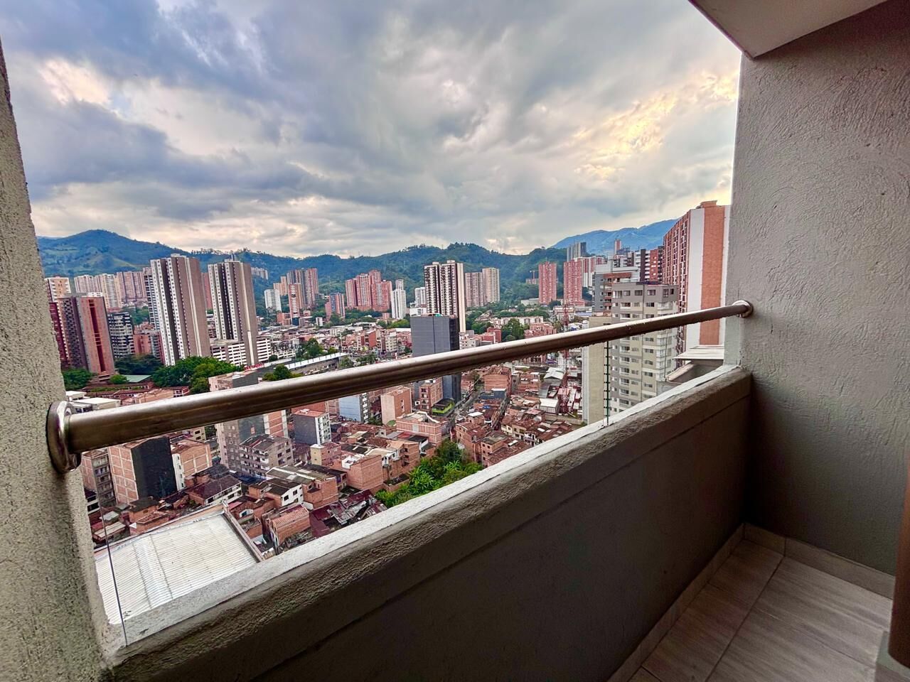 Un balcón con baranda metálica que da a un paisaje urbano con diversos edificios y una montaña al fondo.