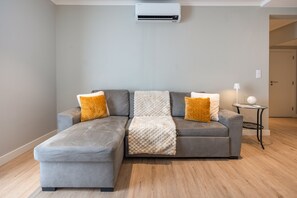En grå hjørnesofa med et pled og mønstrede puder, et lille rundt bord med en lampe, og en hvid aircondition-enhed på væggen.