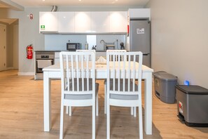 Et moderne køkken med hvide skabe, en mikrobølgeovn og et køleskab. To hvide stole med grå hynder står ved et bord. En brandslukker er monteret på væggen.