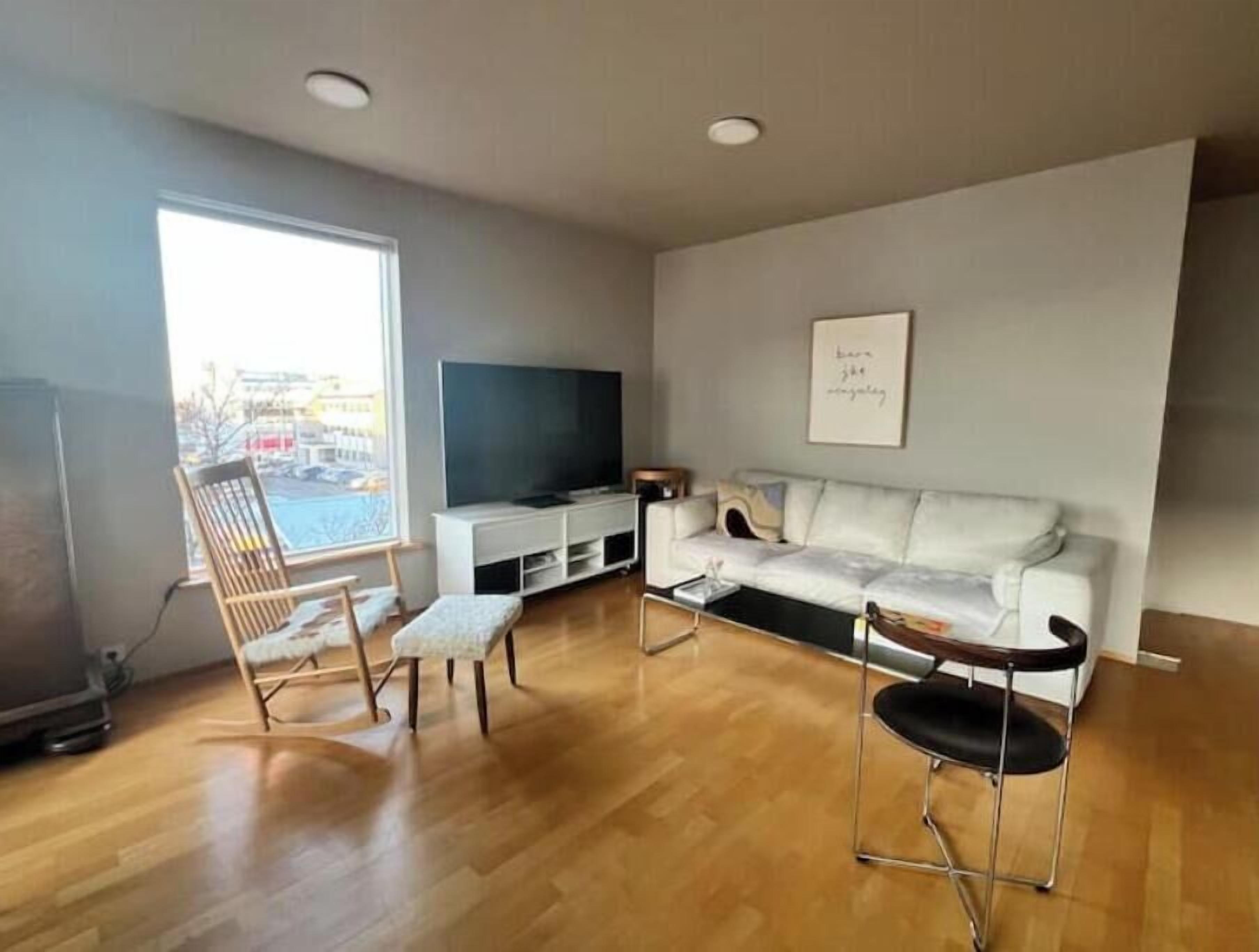 Ein Wohnzimmer mit einem weißen Sofa, einem hölzernen Schaukelstuhl, einem schwarzen Sessel, einem Flachbildfernseher und einem gerahmten Zitat an der Wand.