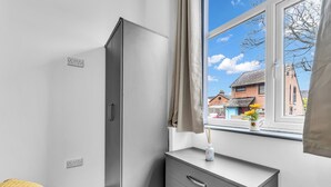 Een kamer met een grijze garderobe, een klein nachtkastje met een bloevas en een raam met uitzicht op gebouwen.