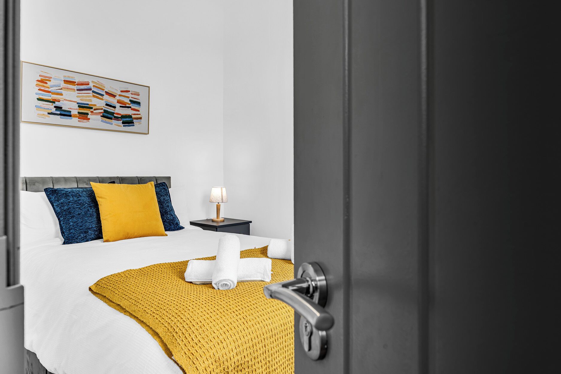 Una habitación de hotel con cama, mesita de noche, lámpara y un cuadro en la pared.