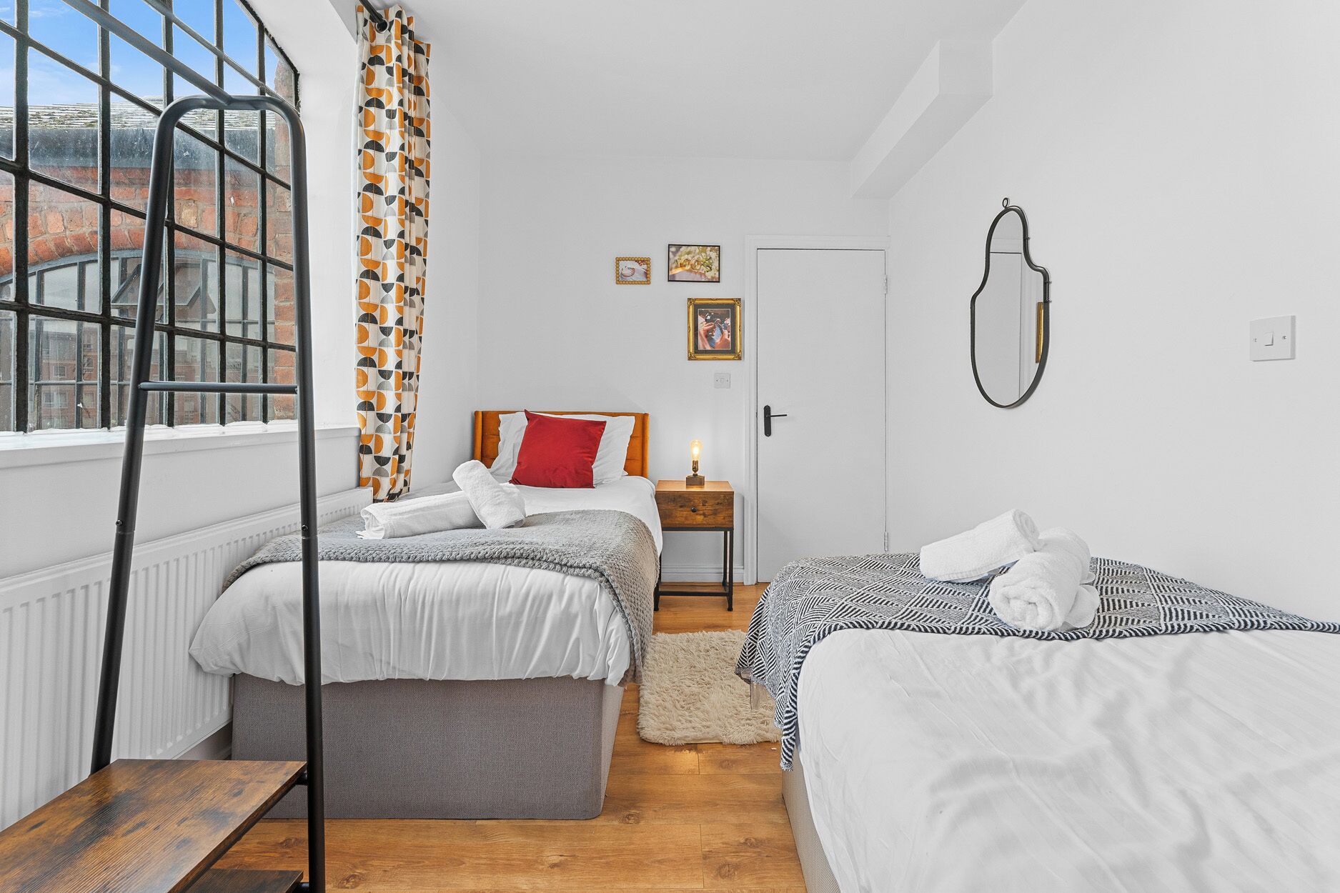 A hotel room with two beds, a window with a view, a radiator, a wooden bench, and a nightstand with a lamp.