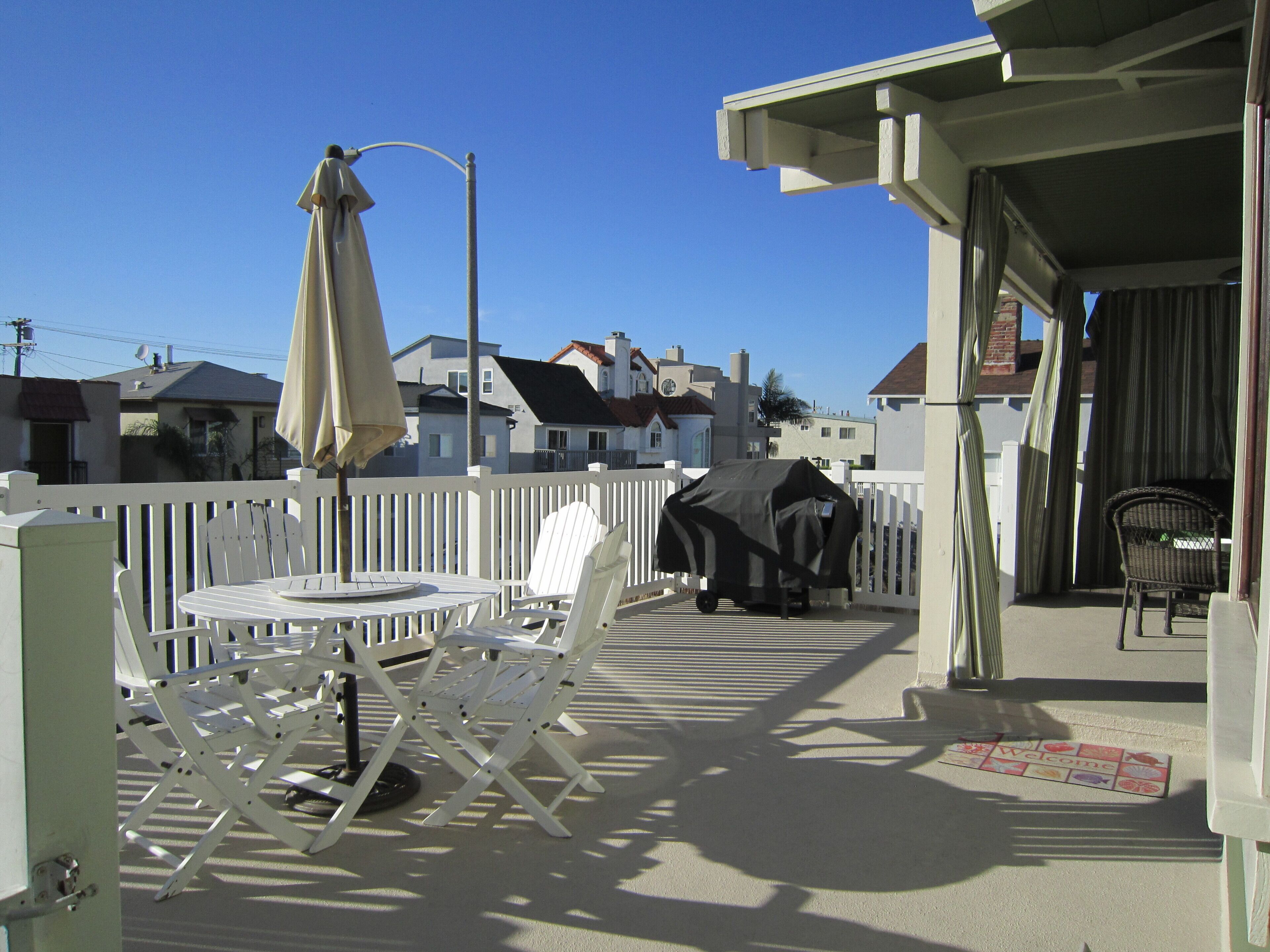 A balcony with a table and chairs, a covered grill, and a view of houses in the background.