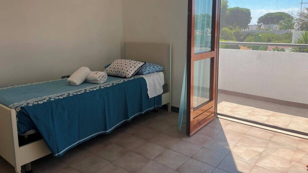 Ein Einzelbett mit blauer Bettdecke und Kissen, eine weiße Wand und Blick auf Grün durch eine Glastür.