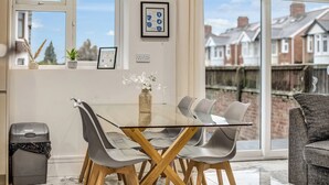 A dining area with a glass table, grey chairs, a trash bin, and a view of houses through large windows. - Oxford City Stay Cowley House (Oxford)