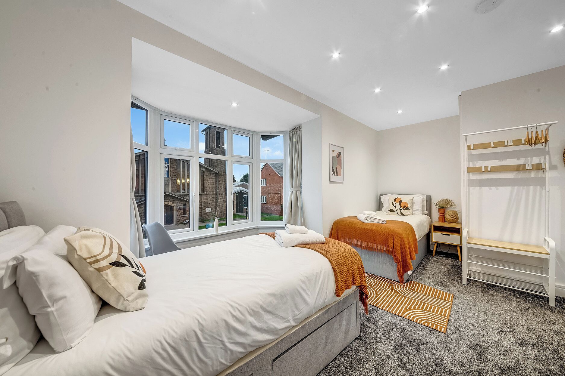 A bedroom with two beds, a window with a view of a brick building, a bedside table with a lamp, and a wardrobe with hanging items.