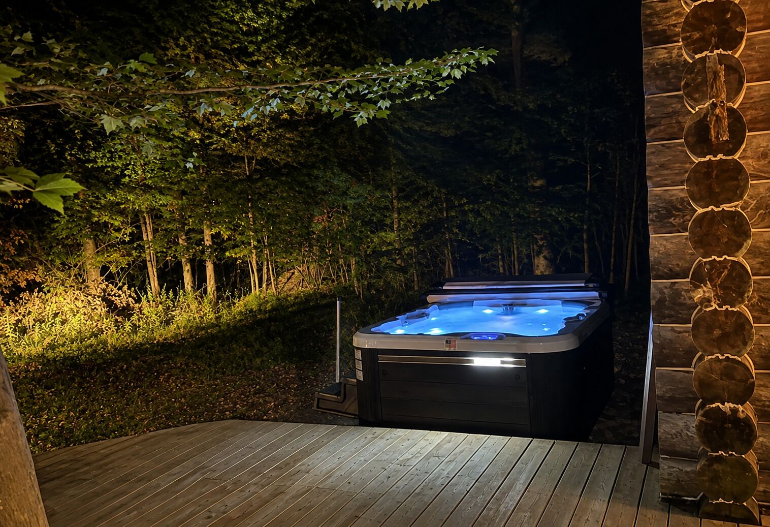 A hot tub in a wooded area with a wooden deck and stone wall.