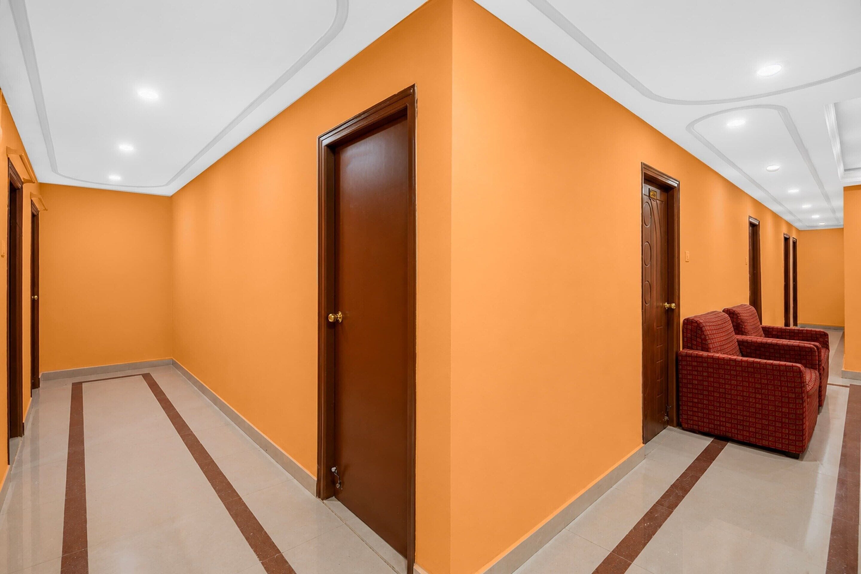A hotel corridor with orange walls, wooden doors, and a seating area with red upholstered chairs.