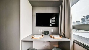 Habitación compacta con una mesa de comedor dispuesta para dos personas, un televisor de pantalla plana montado arriba y vistas de la ciudad a través de una ventana.