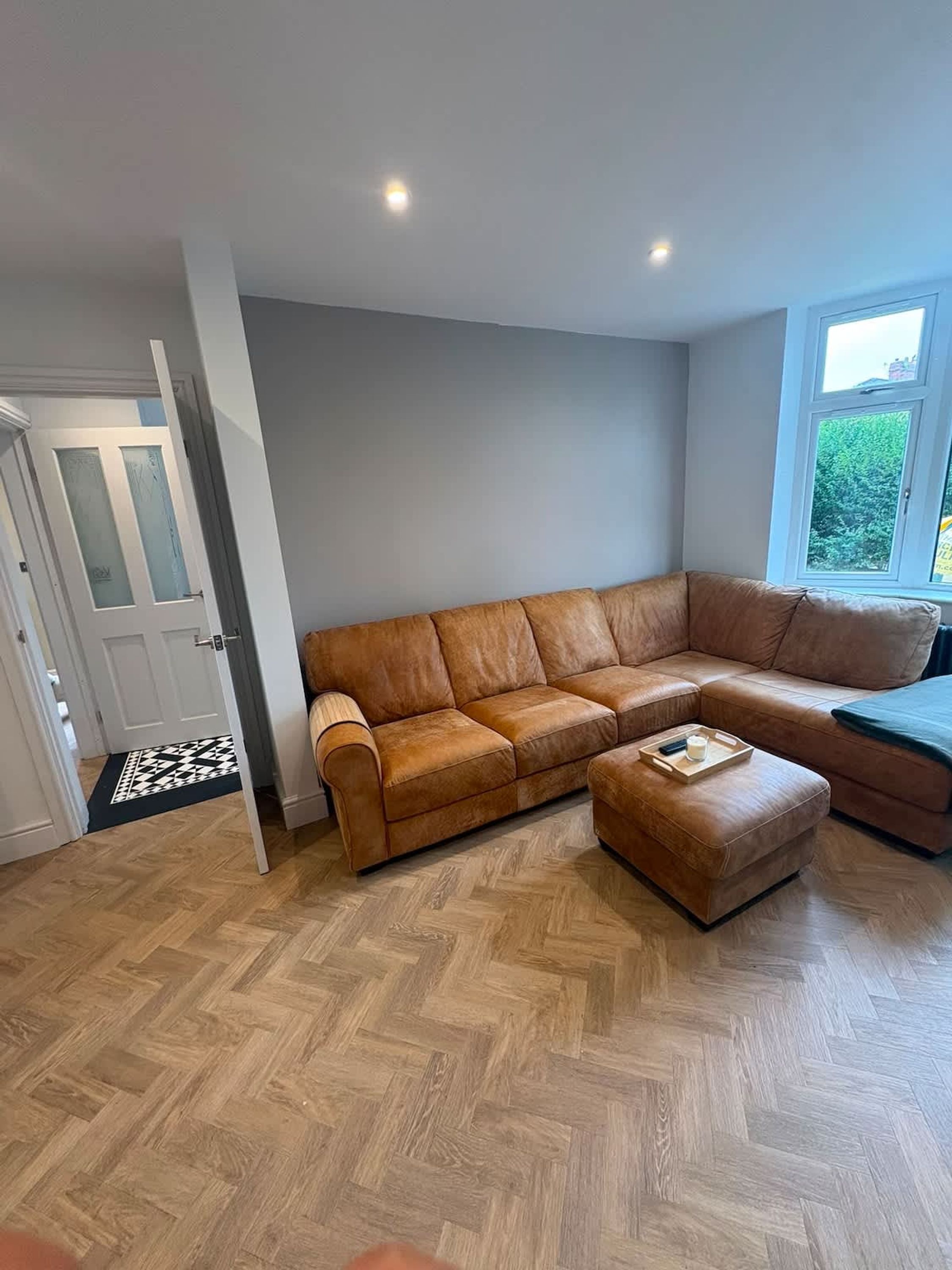 A living room with a brown leather sofa, a matching ottoman, and a herringbone wooden floor.