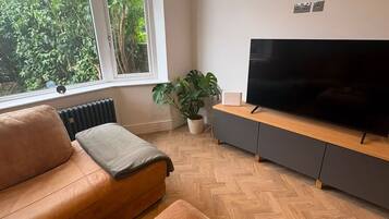 Uma sala de estar com um sofá de couro, uma mesa de centro de madeira com um controle remoto e um copo, uma TV sobre um suporte cinza, e uma janela com vista para o verde.
