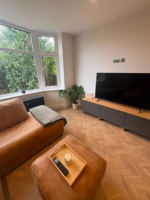 Uma sala de estar com um sofá de couro, uma mesa de centro de madeira com um controle remoto e um copo, uma TV sobre um suporte cinza, e uma janela com vista para o verde.