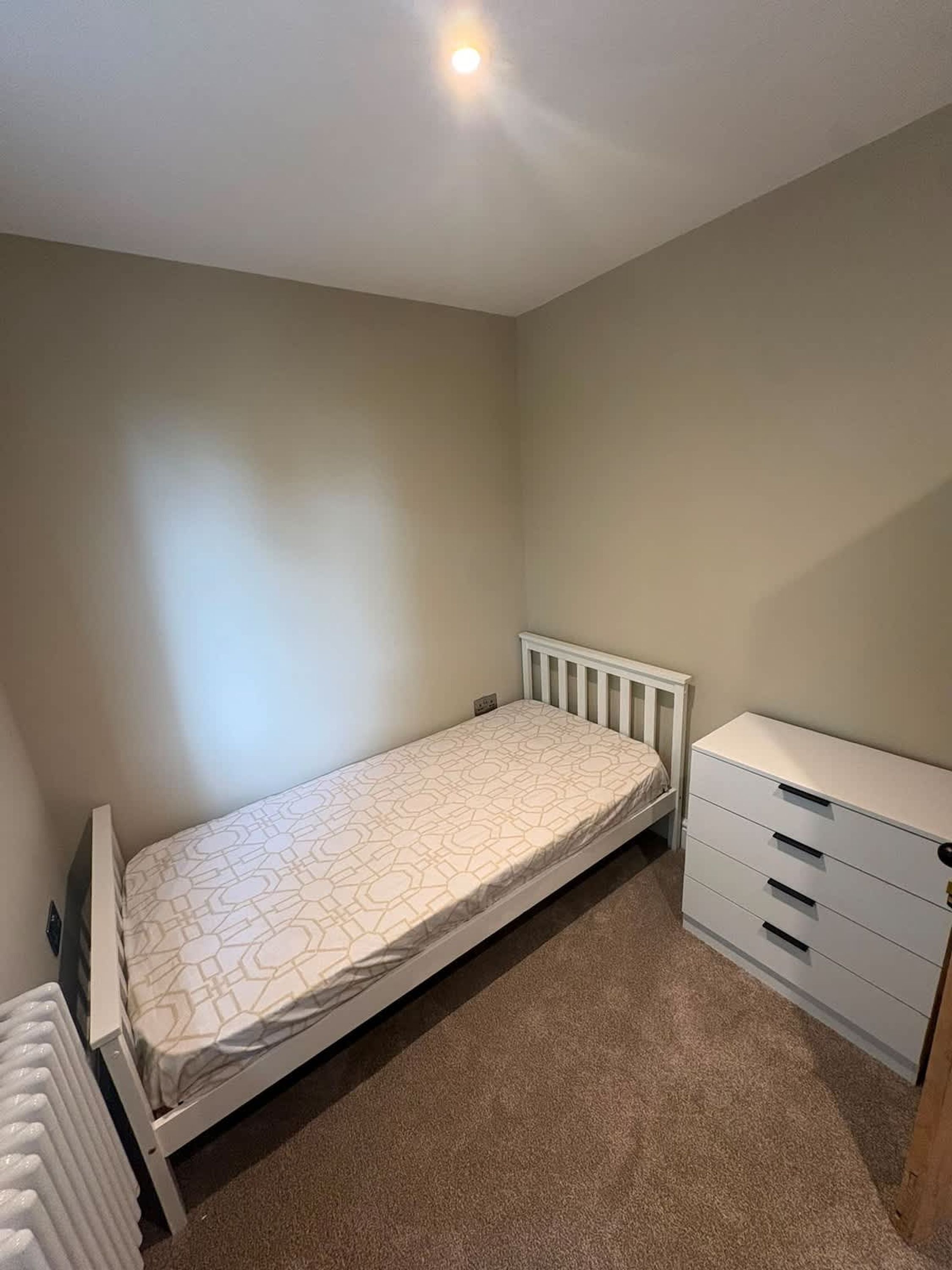 A small, single-bed room with a white radiator, a white dresser with drawers, and a plain wall.