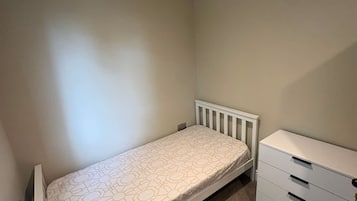 Quarto pequeno com uma cama de solteiro, radiador branco, cômoda branca com gavetas e parede sem detalhes.