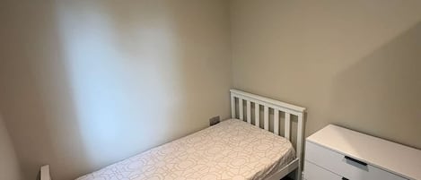 Une petite chambre avec un lit simple, un radiateur blanc, une commode blanche avec des tiroirs et un mur uni.