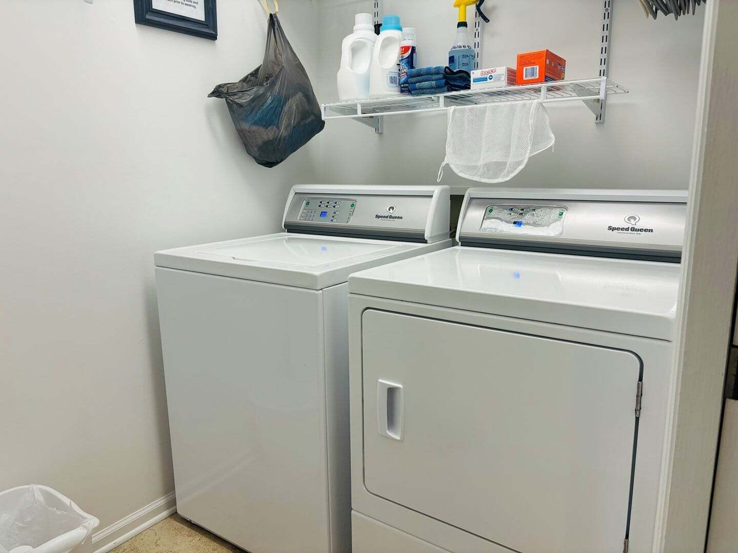 A laundry room with two Speed Queen washers and dryers, a shelf with cleaning supplies, and a trash bin.