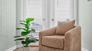 A room with a beige armchair, a potted plant, and white-framed windows. - Sea Monkeys Beach Cottage: 3BR, 2BA  retreat in Holden Beach. (Holden Beach)