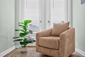 A room with a beige armchair, a potted plant, and white-framed windows. - Sea Monkeys Beach Cottage: 3BR, 2BA  retreat in Holden Beach. (Holden Beach)