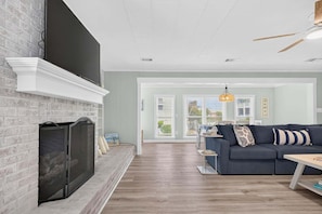 A living room with a fireplace, a blue sofa, and a dining area with a table and chairs. - Sea Monkeys Beach Cottage: 3BR, 2BA retreat in Holden Beach. (Holden Beach)