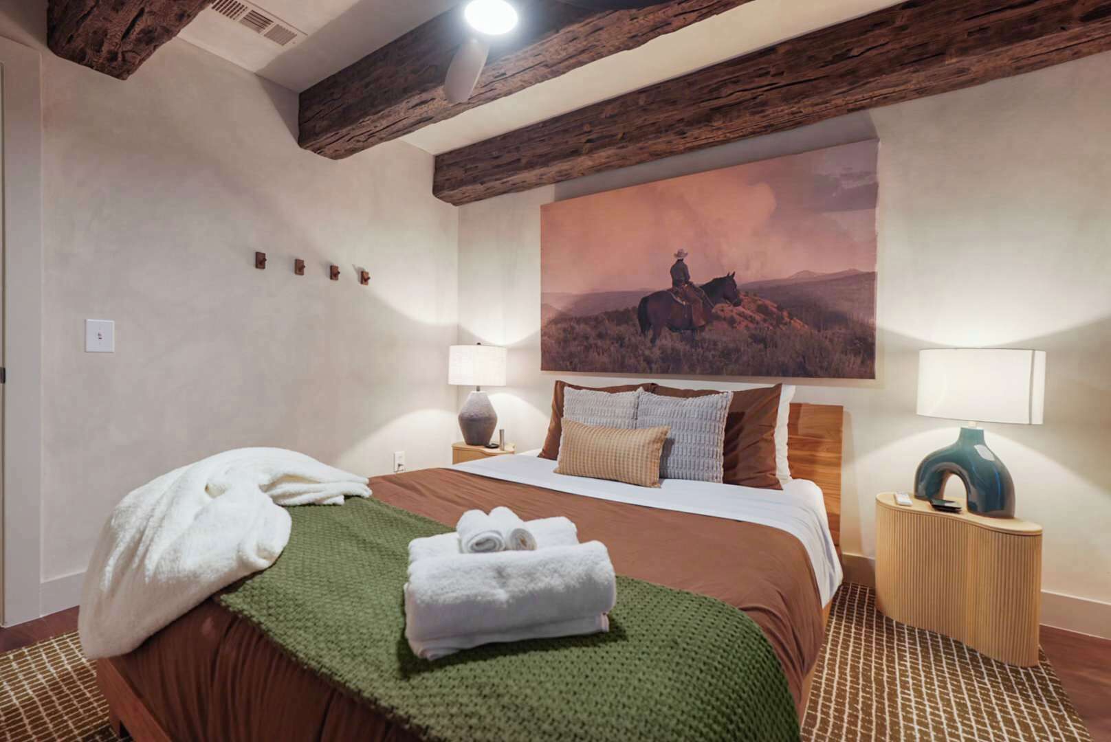 Habitación de hotel con una cama grande, dos lámparas de noche, un cuadro de un caballo en una colina y una viga de techo de madera.