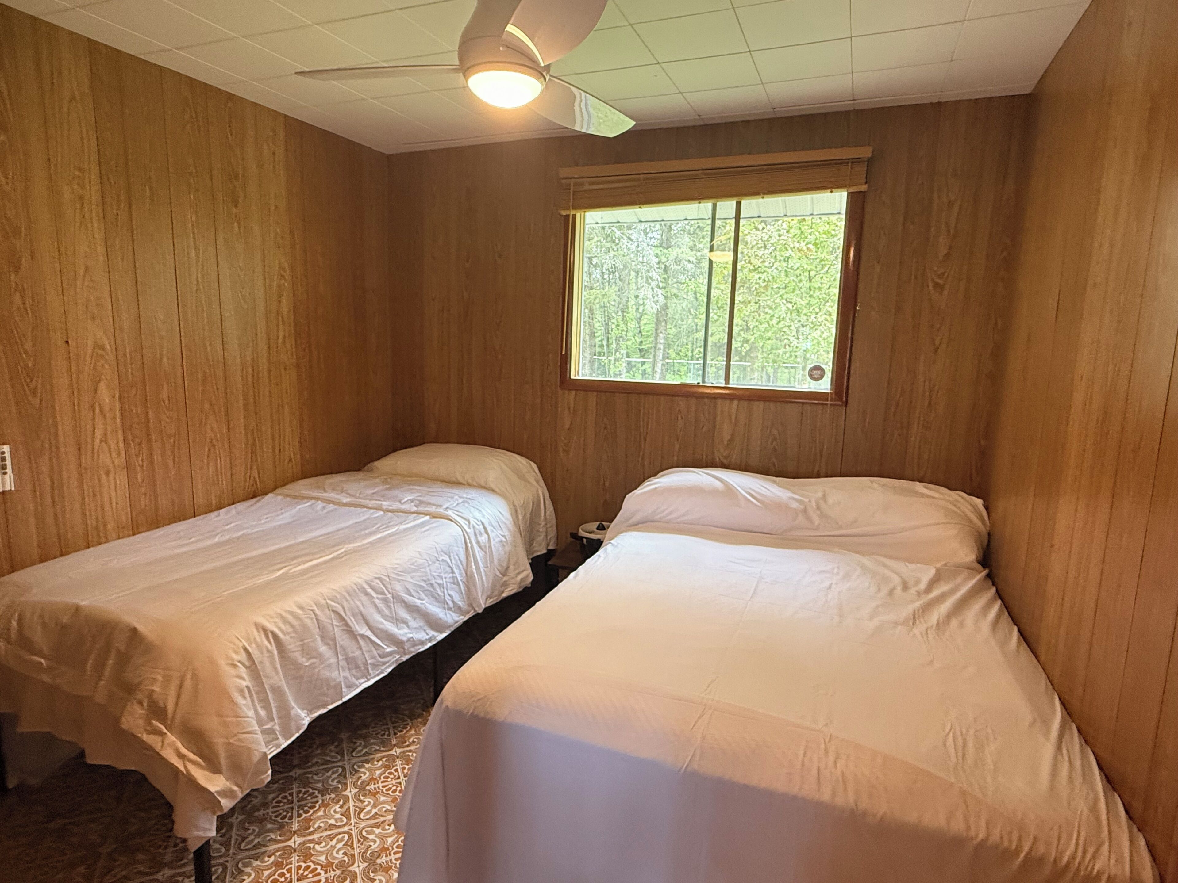 A room with two single beds, wooden walls, a ceiling fan, and a window with a view of trees.