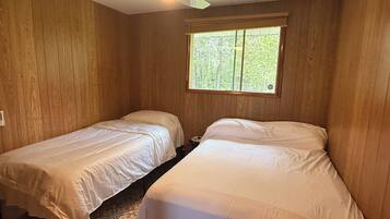 A room with two single beds, wooden walls, a ceiling fan, and a window with a view of trees.