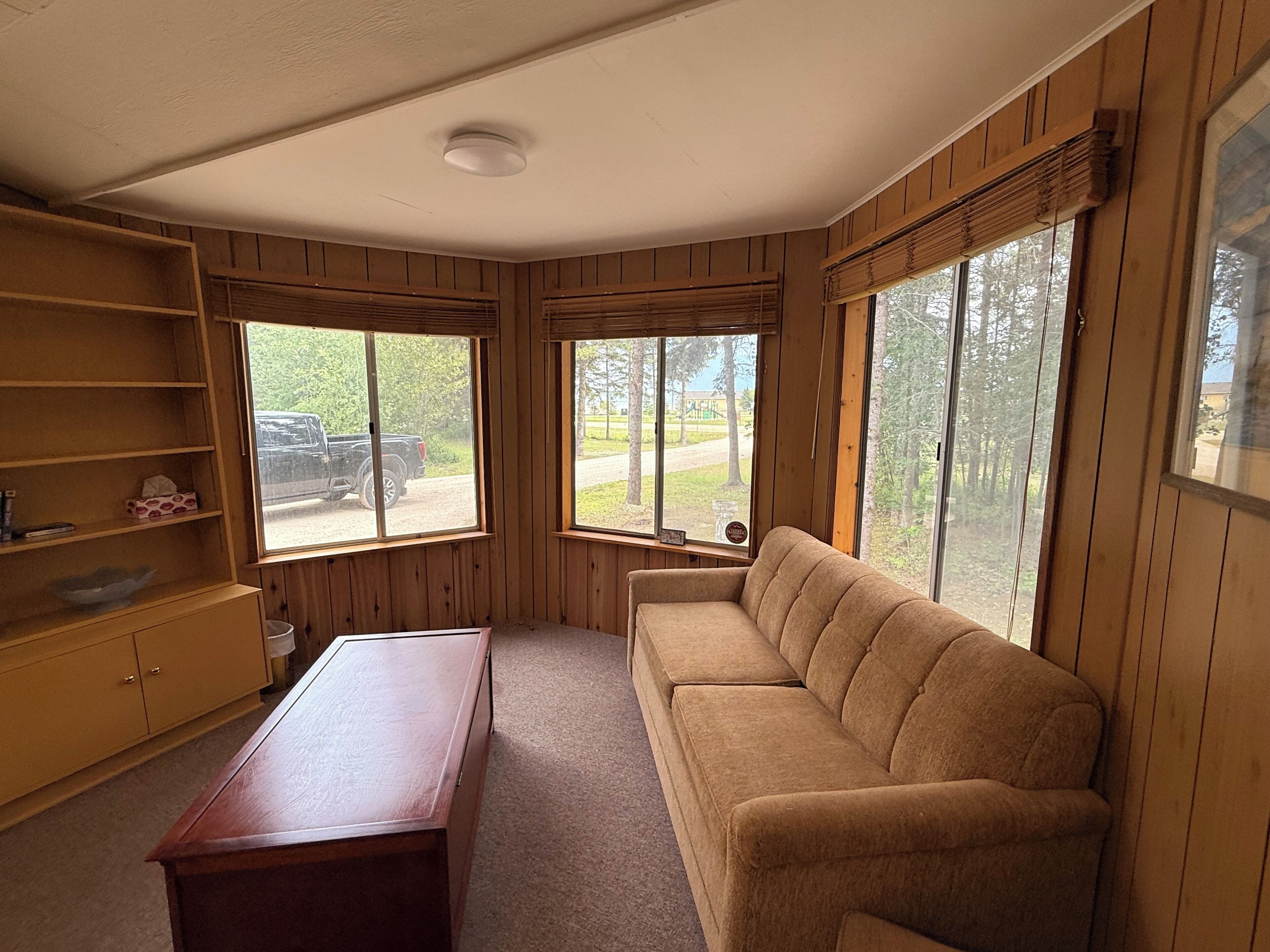 A room with a wooden floor, a beige sofa, a wooden table, and two windows with blinds.