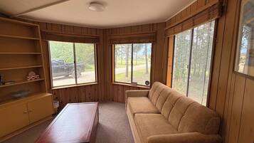 A room with a wooden floor, a beige sofa, a wooden table, and two windows with blinds.
