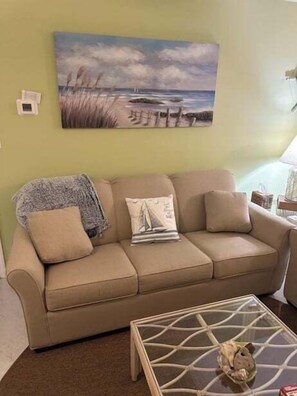 A living room with a beige sofa, a glass coffee table, and a painting of a beach scene on the wall. - Canal-front 3BR, 2BA retreat with dock, WiFi, grill, and dog-friendly comfort. (Supply)