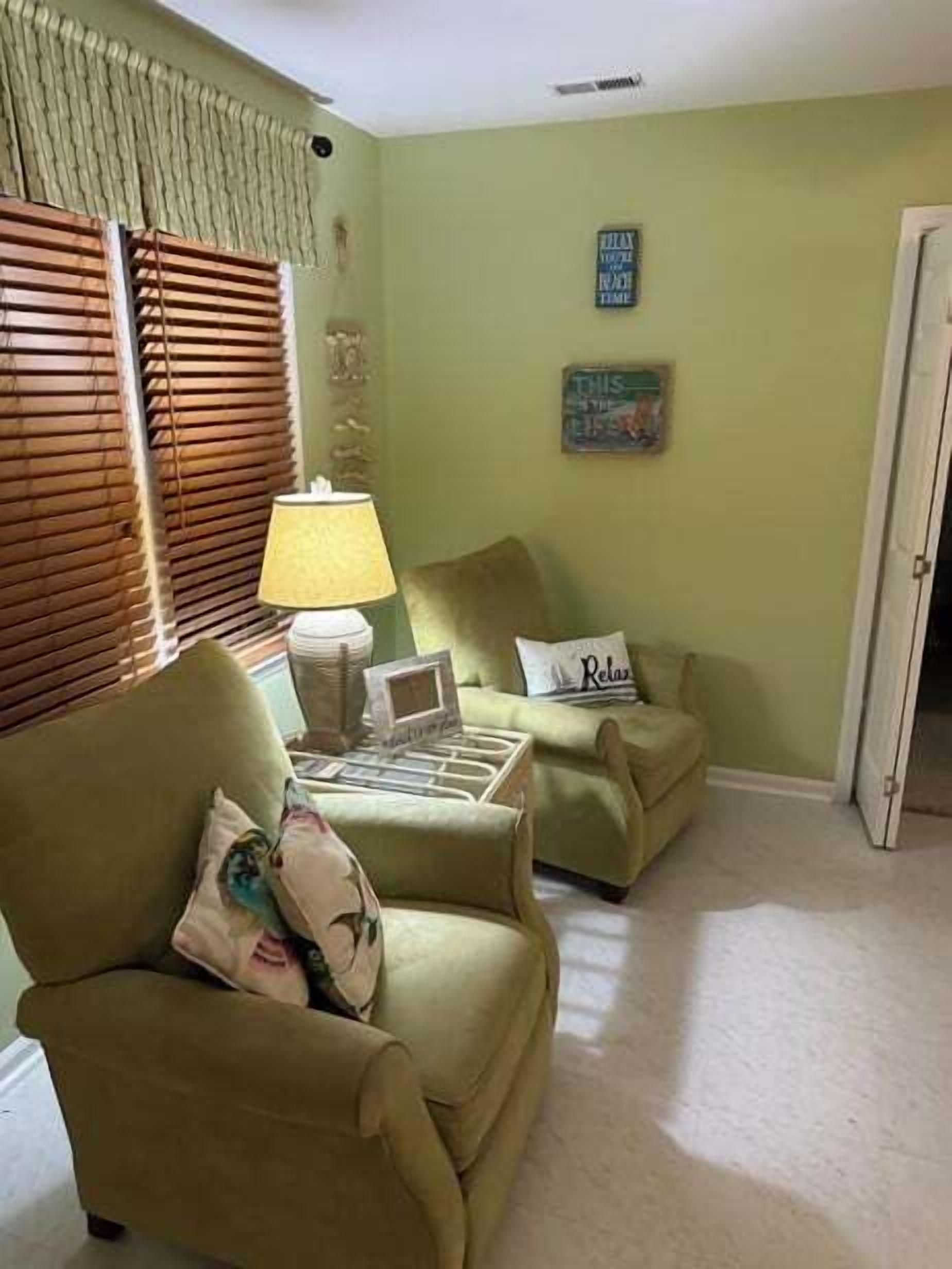 A room with two armchairs, a small table, and a lamp. There is a door on the right and a window with blinds on the left.