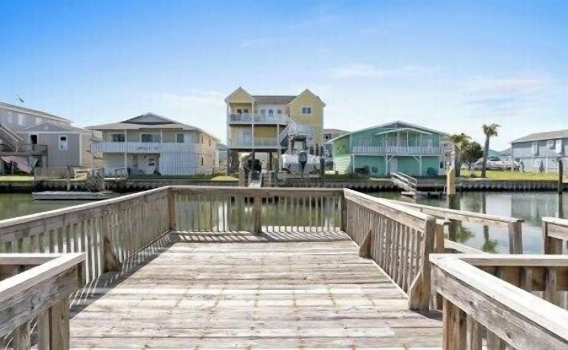 A wooden pier leading to a waterfront property with multiple buildings, including a two-story house with a balcony.