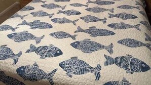 A bed with a fish pattern bedspread, a starfish pillow, a framed picture on the wall, and a nightstand with a lamp and a decorative item. - Canal-front 3BR, 2BA retreat with dock, WiFi, grill, and dog-friendly comfort. (Supply)