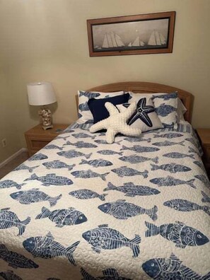 Ein Bett mit Fischmuster-Bettdecke, ein Meeresschnecken-Kissen, ein gerahmtes Bild an der Wand und ein Nachttisch mit Lampe und Dekoration.